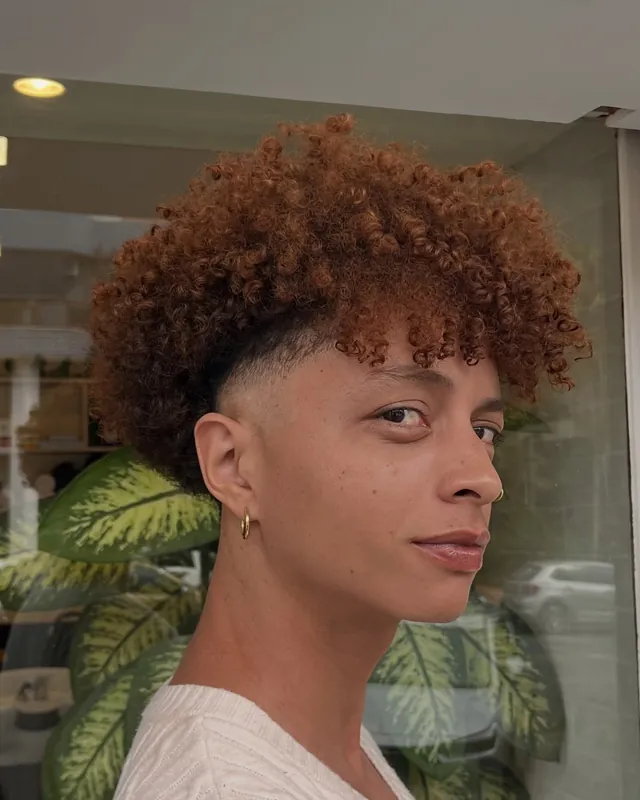 Cabelo cacheado cor de cobre, com degradê lateral, visto de perfil, num ambiente com plantas