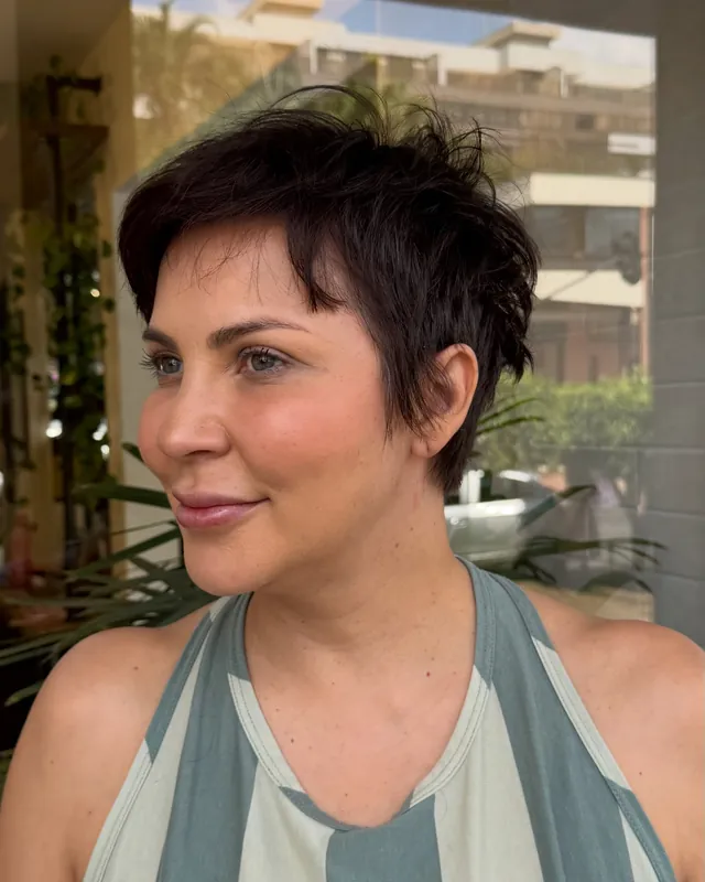 Mulher de corte pixie, sorrindo, com o reflexo da rua e das plantas do salão ao fundo